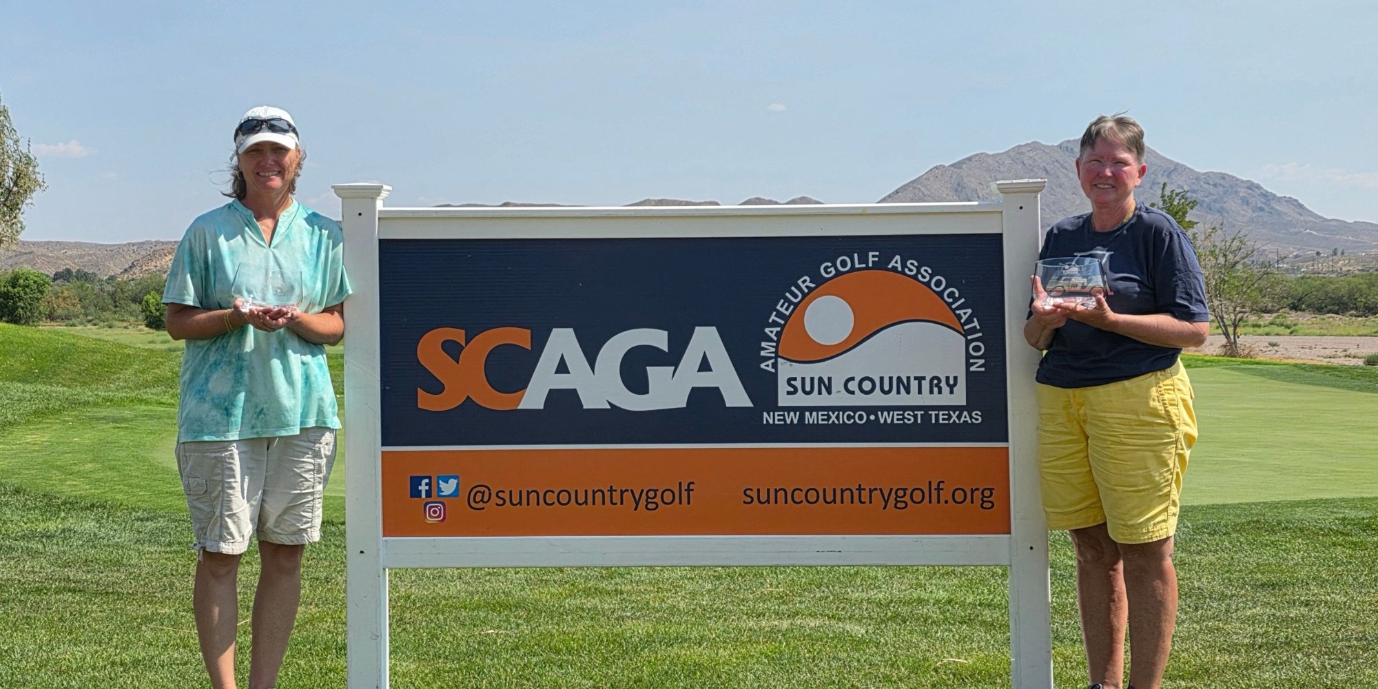 Two SCAGA champions with their trophies beside the SCAGA sign